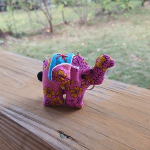 May include: A pink fabric elephant keychain with a floral pattern and a blue and white patterned saddle. The elephant's trunk is raised.