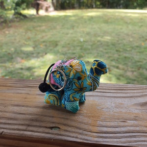May include: A blue, yellow, and pink patterned elephant keychain with a black tassel. The elephant is made of fabric and has a silver key ring.