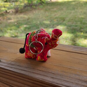 May include: A red and white fabric elephant keychain with a green and gold patterned design. The elephant has a small black tail with a ball at the end. The keychain has a silver ring.