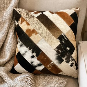May include: A square throw pillow featuring a patchwork design of cowhide strips arranged in a diagonal pattern. The hide pieces display a variety of natural colors including black, white, tan, and dark brown, with some sections showing spotted textures.