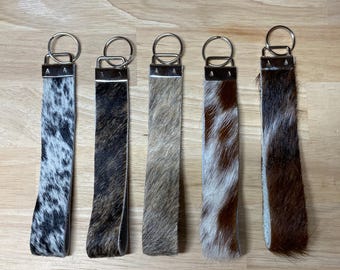 May include: Five wristlet keychains made of cowhide leather with varied patterns including black and white spots, dark brown, light tan, brown and white patches, and dark brown and black stripes, each featuring a silver metal clasp and ring.