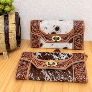 May include: Two rectangular wallets featuring brown and white cowhide panels framed by tan leather with intricate floral tooling. Each wallet has a central brass-toned twist lock closure and small turquoise accents embedded within the floral patterns.