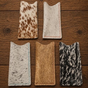 May include: A collection of five leather card holders with a unique cowhide pattern. The holders vary in color, including brown and white, gray, and black and white. Each holder has a stitched edge in a contrasting color.