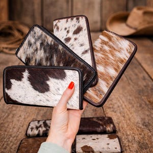 Puede incluir: Una colección de carteras de piel de vaca en varios tonos de marrón, blanco y negro. Las carteras tienen forma rectangular con cierre de cremallera y ribete de cuero marrón oscuro. Algunas carteras se sostienen en una mano con esmalte de uñas rojo.