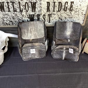 Puede incluir: Dos mochilas confeccionadas en piel de vaca gris y marrón con textura natural de pelo. Cada bolsa cuenta con un compartimento principal grande con cierre de solapa, un bolsillo frontal con cremallera más pequeño y una etiqueta blanca pequeña.