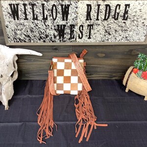 May include: A square crossbody bag featuring a brown and white checkered cowhide pattern on the front. The bag includes a long, tooled leather strap and long brown suede fringe hanging from both sides of the bag, with a zipper closure at the top.