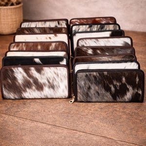 May include: A collection of cowhide wallets with various patterns and colors, including black, white, and brown. Each wallet has a zipper closure and a rectangular shape. The wallets are arranged on a wooden surface.
