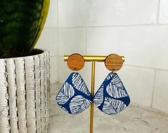 May include: A pair of blue and white patterned dangle earrings with wooden tops. The earrings are displayed on a gold stand.