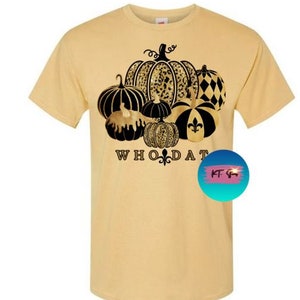 May include: A light yellow t-shirt featuring a graphic of several pumpkins in various patterns and colors, including black, gold, and white. The text "WHO DAT" is printed below the pumpkins. The shirt has short sleeves and a crew neck.