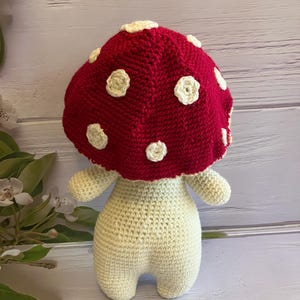 May include: A crocheted mushroom toy with a red cap and white spots. The mushroom has a white body and is standing on a light wood surface.