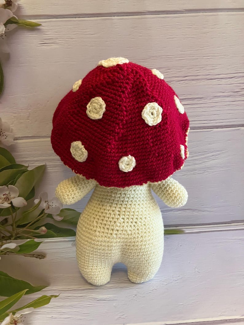 May include: A crocheted mushroom toy with a red cap and white spots. The mushroom has a white body and is standing on a light wood surface.