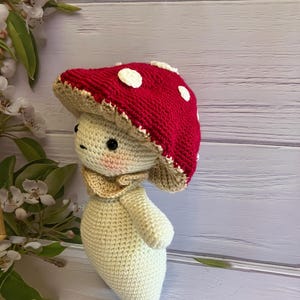 May include: A crocheted mushroom toy with a red and white cap and a cream-colored body. The toy is standing on a light wood surface.