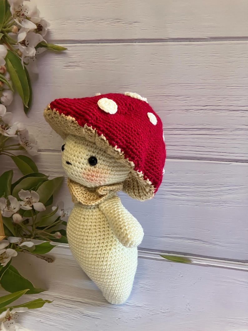 May include: A crocheted mushroom toy with a red and white cap and a cream-colored body. The toy is standing on a light wood surface.