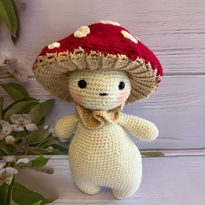 May include: A crocheted mushroom toy with a red and white cap and a cream-colored body. The toy has a cute face with black eyes and pink cheeks.