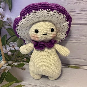 May include: A crocheted mushroom-shaped plush toy with a white body, purple hat, and a purple bow. The toy has black eyes and a pink blush.