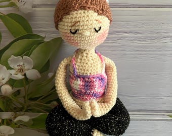 May include: A crocheted doll with brown hair, closed eyes, and a pink and purple top, sitting in a yoga pose wearing black pants with gold glitter.