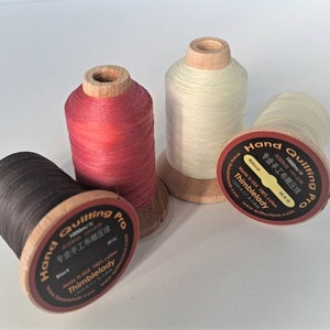May include: Four spools of thread for hand quilting. The spools are made of wood and have labels that say "Hand Quilting Pro" and "Thimblelady". The thread colours are black, red, white, and natural.