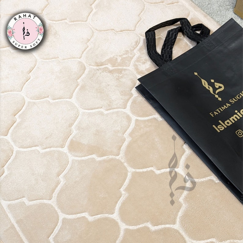 Puede incluir: Una alfombra de oraci&oacute;n beige con un patr&oacute;n geom&eacute;trico. La alfombra est&aacute; hecha de tela suave y felpa y tiene una bolsa de compras negra con el texto "Fatima Sugha Islamic" en ella.