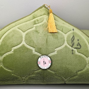 Puede incluir: Una alfombra de oraci&oacute;n verde con un patr&oacute;n geom&eacute;trico y una borla. La alfombra tiene un logotipo circular con el texto "RAHAT SUPER SOFT" y caligraf&iacute;a &aacute;rabe.