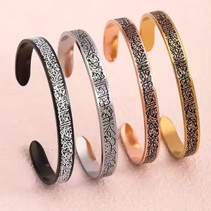May include: Four open-ended cuff bracelets in black, silver, rose gold, and gold. Each bracelet features an engraved design in a contrasting color. The bracelets are displayed on a light pink surface.