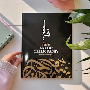 Op de afbeelding: Een zwart boek getiteld Learn Arabic Calligraphy door Fatima Sughra met witte tekst en een goudkleurig Arabisch kalligrafieontwerp onderaan. De kaft heeft een verticaal wit Arabisch teken in het midden en een glanzende afwerking op de voorzijde.
