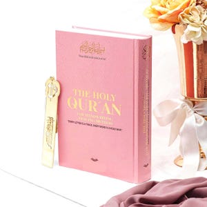 May include: A pink hardback book titled "The Holy Qur'an" with gold lettering and a gold bookmark. The book is for handwriting tracing and includes the phrase "Every letter is a trace, every word is a road map."