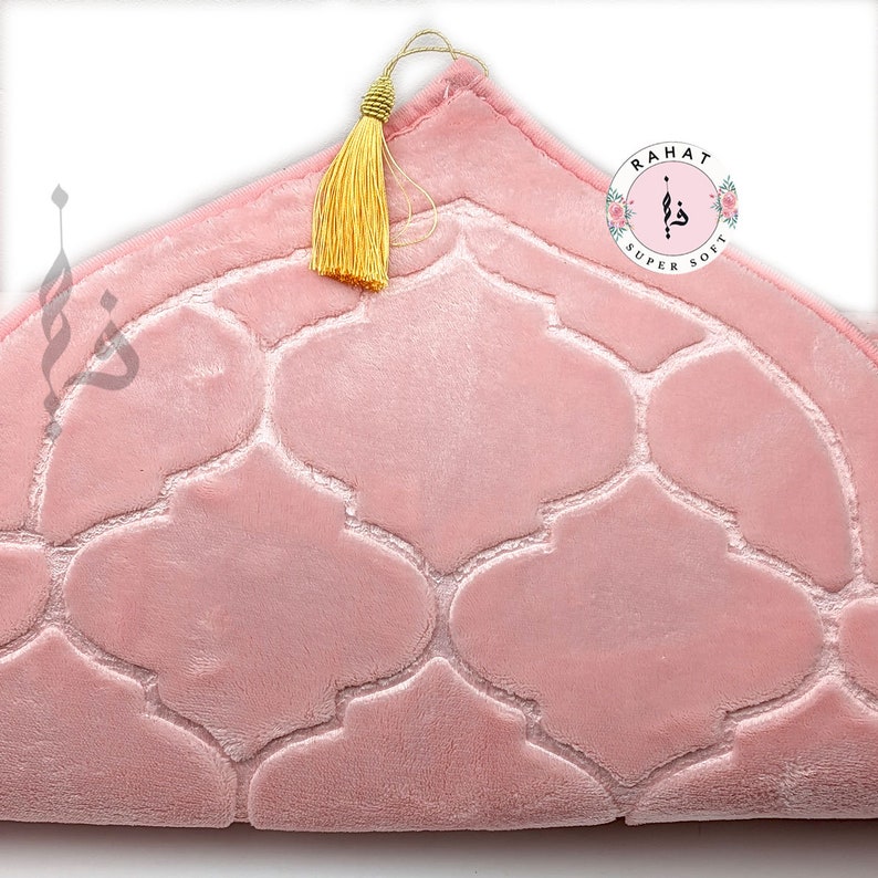 Puede incluir: Una alfombra de oraci&oacute;n rosa con un patr&oacute;n geom&eacute;trico y una borla dorada. La alfombra est&aacute; etiquetada como "Rahat Super Soft".