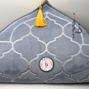 Puede incluir: Una alfombra de oraci&oacute;n gris con un patr&oacute;n geom&eacute;trico blanco y una borla amarilla. La alfombra tiene un logotipo circular con el texto "RAHAT SUPER SOFT" en blanco.