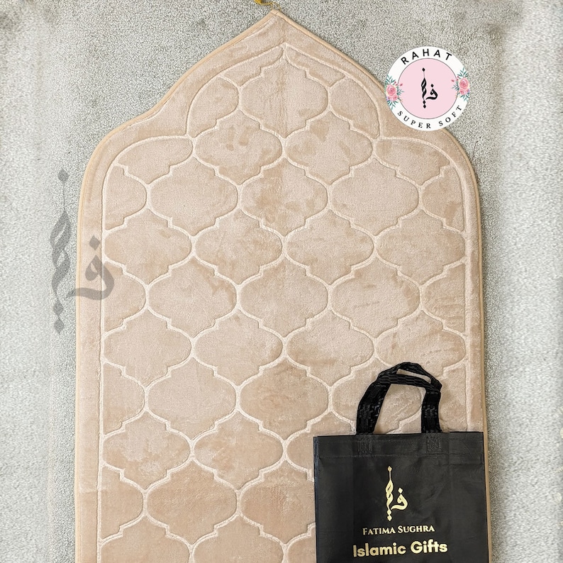 Puede incluir: Una alfombra de oraci&oacute;n beige con un patr&oacute;n geom&eacute;trico y una bolsa de mano negra con el texto "Fatima Sughra Islamic Gifts" y un logotipo. La bolsa de mano tiene un asa negra.
