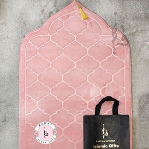 Puede incluir: Una alfombra de oraci&oacute;n rosa con un dise&ntilde;o acolchado y una borla dorada. La alfombra est&aacute; empaquetada en una bolsa de mano negra con el texto "Fatima Sughra Islamic Gifts".