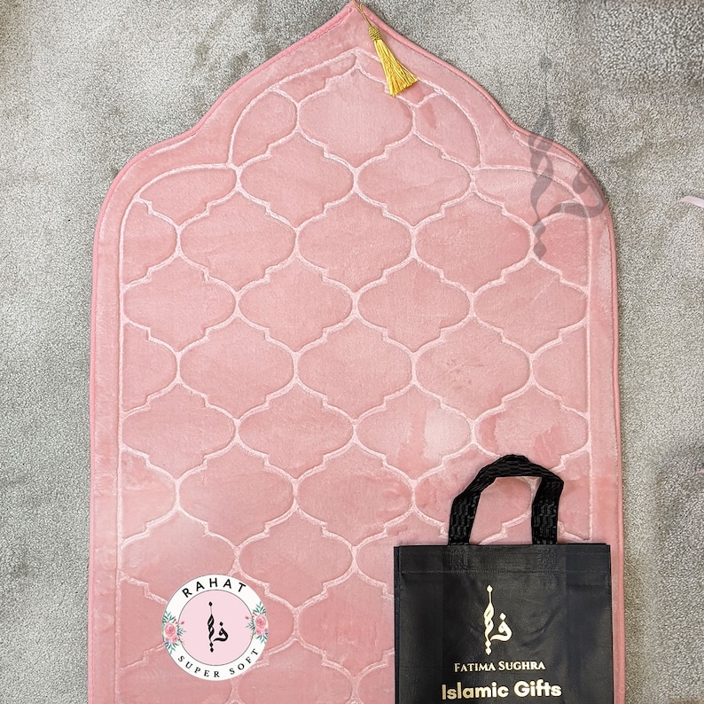 Puede incluir: Una alfombra de oraci&oacute;n rosa con un dise&ntilde;o acolchado y una borla dorada. La alfombra est&aacute; empaquetada en una bolsa de mano negra con el texto "Fatima Sughra Islamic Gifts".