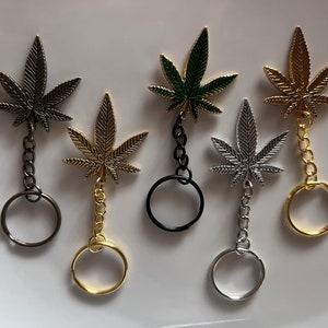 Puede incluir: Cinco llaveros con diferentes dijes de hoja de marihuana de metal de colores. Los dijes están unidos a llaveros plateados, dorados y negros.