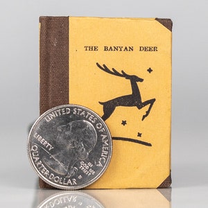 May include: A small, yellow book with a brown spine. The cover features a black silhouette of a deer jumping with stars in the background. The title "THE BANYAN DEER" is printed on the cover. A US quarter is placed in front of the book for scale.
