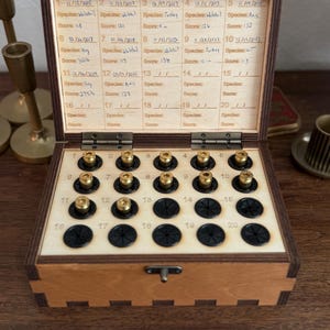 May include: A wooden storage box with a hinged lid featuring a printed grid for tracking twenty entries, including dates, species, and scores. The interior base contains twenty circular slots, with thirteen brass casings inserted into the first thirteen spots.