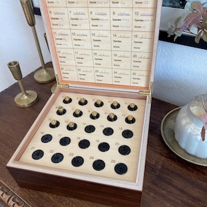May include: A wooden box with a hinged lid, featuring a grid for recording data. The box contains numbered compartments with small, gold-coloured knobs and black circular inserts. The lid has handwritten entries. Two brass candle holders are visible.