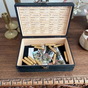 May include: A black wooden keepsake box with a hinged lid, open to reveal a black interior with brass bullet casings and photos. The lid has a calendar-like grid with handwritten entries. The box sits on a wooden table.