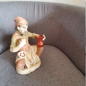 Peut inclure: Une figurine en céramique d'un homme portant un chapeau rouge et tenant une cruche brune. La figurine est assise sur une surface en tissu gris.