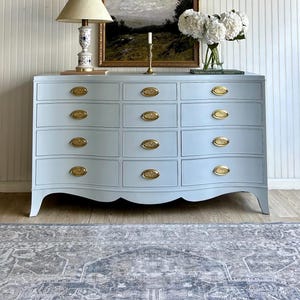 May include: A light blue dresser with gold-toned hardware. The dresser has a curved front with multiple drawers. A lamp, artwork, and flowers are displayed on top. The room has a rug and white paneling.