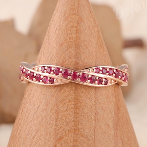 May include: A rose gold band ring featuring a twisted design set with small, round, vibrant pinkish-red gemstones. The ring is displayed on a smooth, light-colored wooden cone stand. The metal has a polished finish and a delicate, interwoven structure.