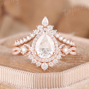 May include: A rose gold ring set featuring a pear-shaped center stone surrounded by a halo of small round stones. The band has a twisted design with additional round stones. The text DejavuJewelryCo is visible in the upper corners of the image.