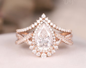 Puede incluir: Un juego de anillos de compromiso de oro rosa con una piedra central en forma de pera rodeada por un halo de piedras más pequeñas. La banda tiene un diseño tejido con piedras de acento adicionales, creando un aspecto brillante y elegante.