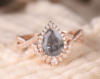 Puede incluir: Un anillo de compromiso de oro rosa con un diamante gris en forma de pera engastado en un halo de diamantes más pequeños. La banda del anillo está retorcida y presenta un diseño delicado.
