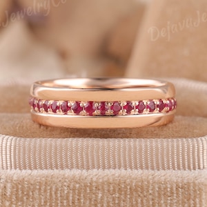May include: A rose gold band ring featuring a central channel set with a continuous row of small, round, vibrant red rubies. The metal band has smooth, polished edges above and below the row of gemstones, creating a clean, symmetrical design.