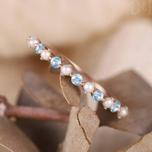 May include: A delicate rose gold ring featuring a row of small, round, light blue gemstones and white pearls. The ring is set against a soft, neutral background, highlighting the jewelry's elegant design.