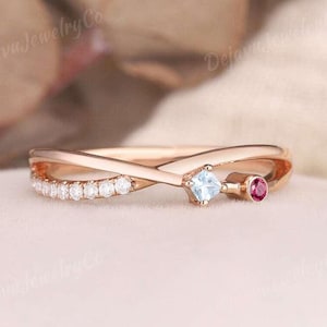 May include: Rose gold ring with a crisscross design. The ring features a row of small white stones, a square light blue stone, and a small round red stone. The ring is set on a light pink surface.