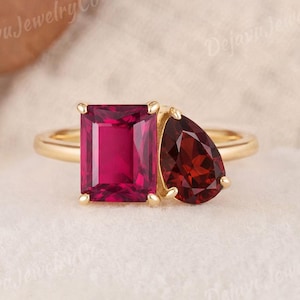 May include: A gold ring featuring a large, rectangular ruby and a smaller, pear-shaped garnet. The gemstones are a deep red color, set side-by-side on a polished gold band. The ring is a statement piece, perfect for special occasions.