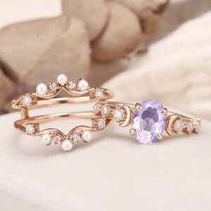 May include: A set of two rose gold rings with a delicate design. The larger ring features a light purple oval gemstone surrounded by small diamonds and crescent moon shapes. The smaller ring has a curved design with small pearls and diamonds.