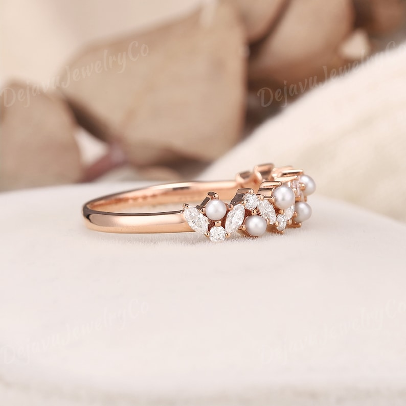 May include: Rose gold ring with a floral design, featuring pearls and marquise-cut diamonds. The ring has a delicate band and a cluster of gemstones. The ring is set on a white surface.