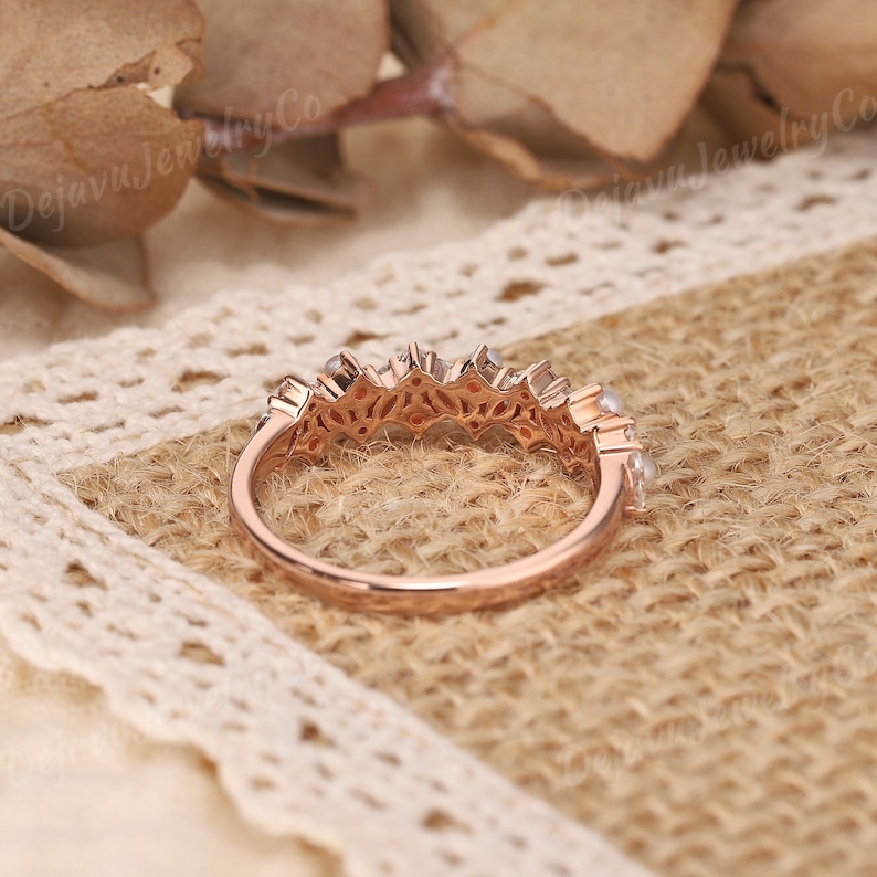 May include: Rose gold ring with an intricate design. The ring has small, clear gemstones set along the top, with an openwork filigree pattern. The ring is displayed on a textured, neutral-toned surface.