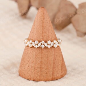 May include: A gold ring with a floral pattern, set with pearls and clear gemstones. The ring is displayed on a wooden cone. The design features alternating pearls and marquise-cut gemstones, showcasing the ring's intricate details and craftsmanship.